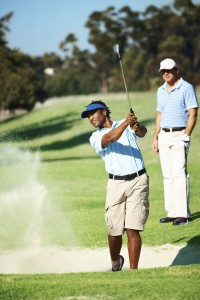 African American golfer swinging from a sand bunker, a friend in the background